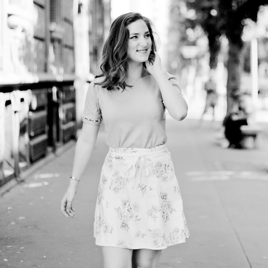 Une jeune femme souriante qui marche dans une rue en plein air, vêtue d'un t-shirt et d'une jupe à motifs floraux, avec des arbres et des bâtiments en arrière-plan.