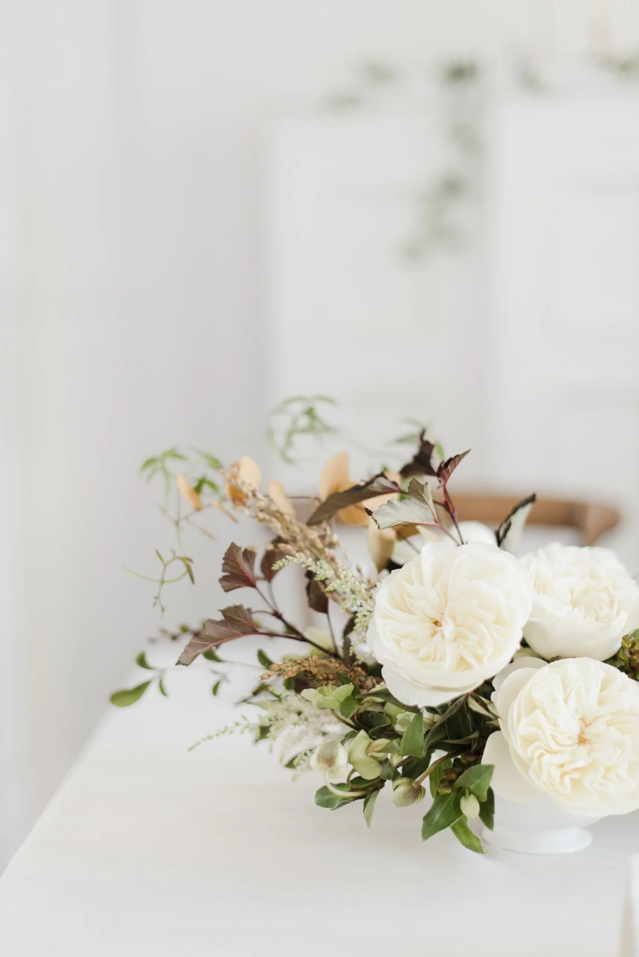 Un arrangement floral composé de roses blanches, de feuillage vert et de branches sèches, placé dans un vase blanc sur une table blanche devant un mur clair.