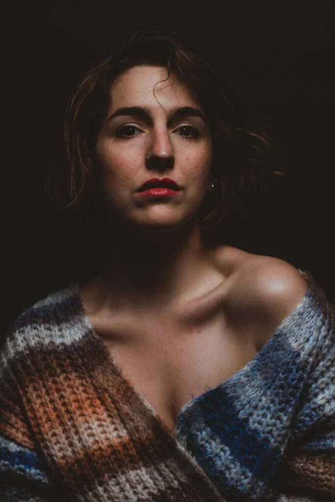 Marie Benquet avec une peau claire, une boucle d'oreille, portant un pull en laine à rayures et un maquillage rouge à lèvres, contre un fond noir.