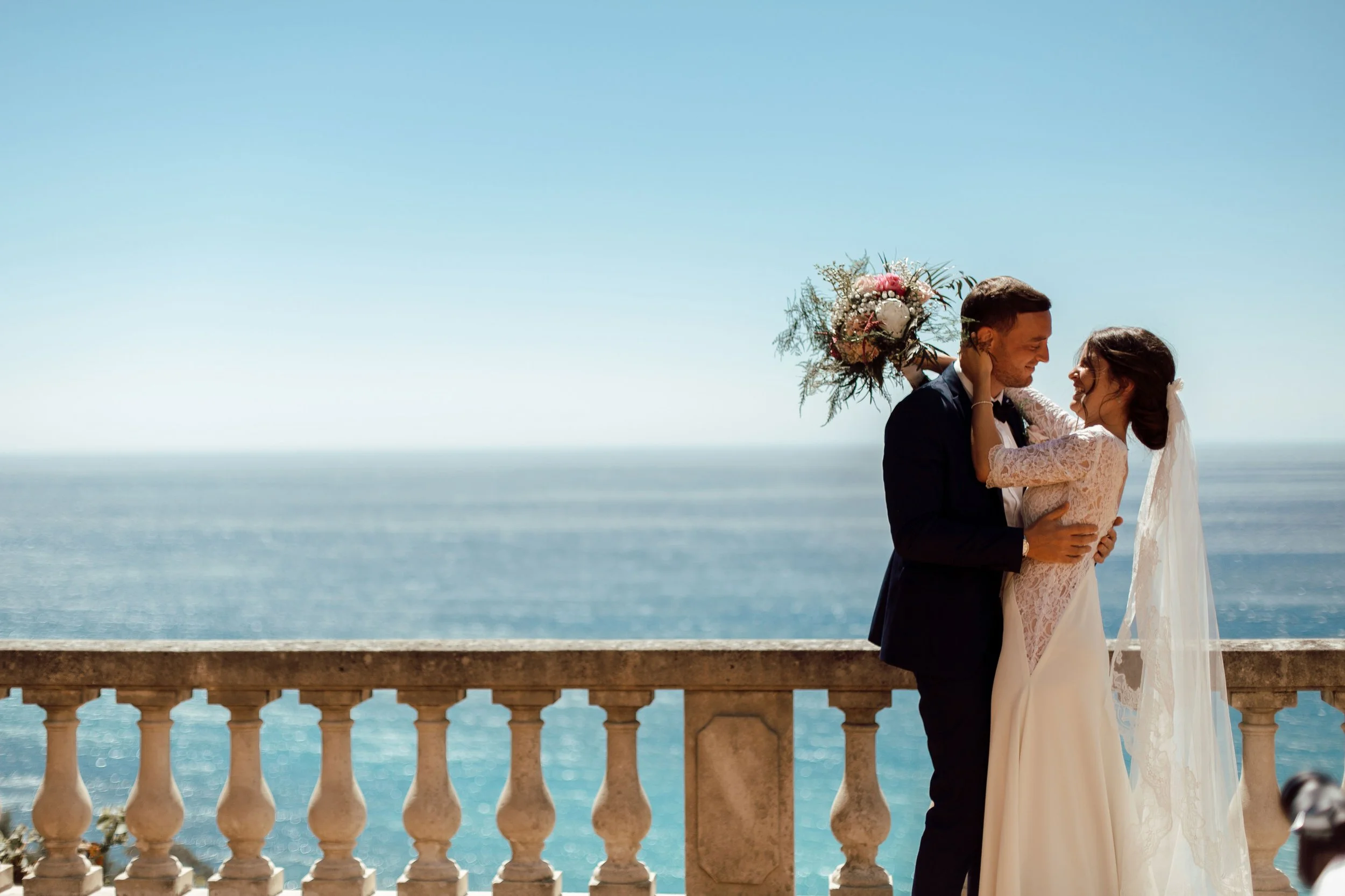 Un couple de mariés s'embrassent sur une terrasse près de la mer, avec une vue sur la mer et le ciel bleu en arrière-plan.