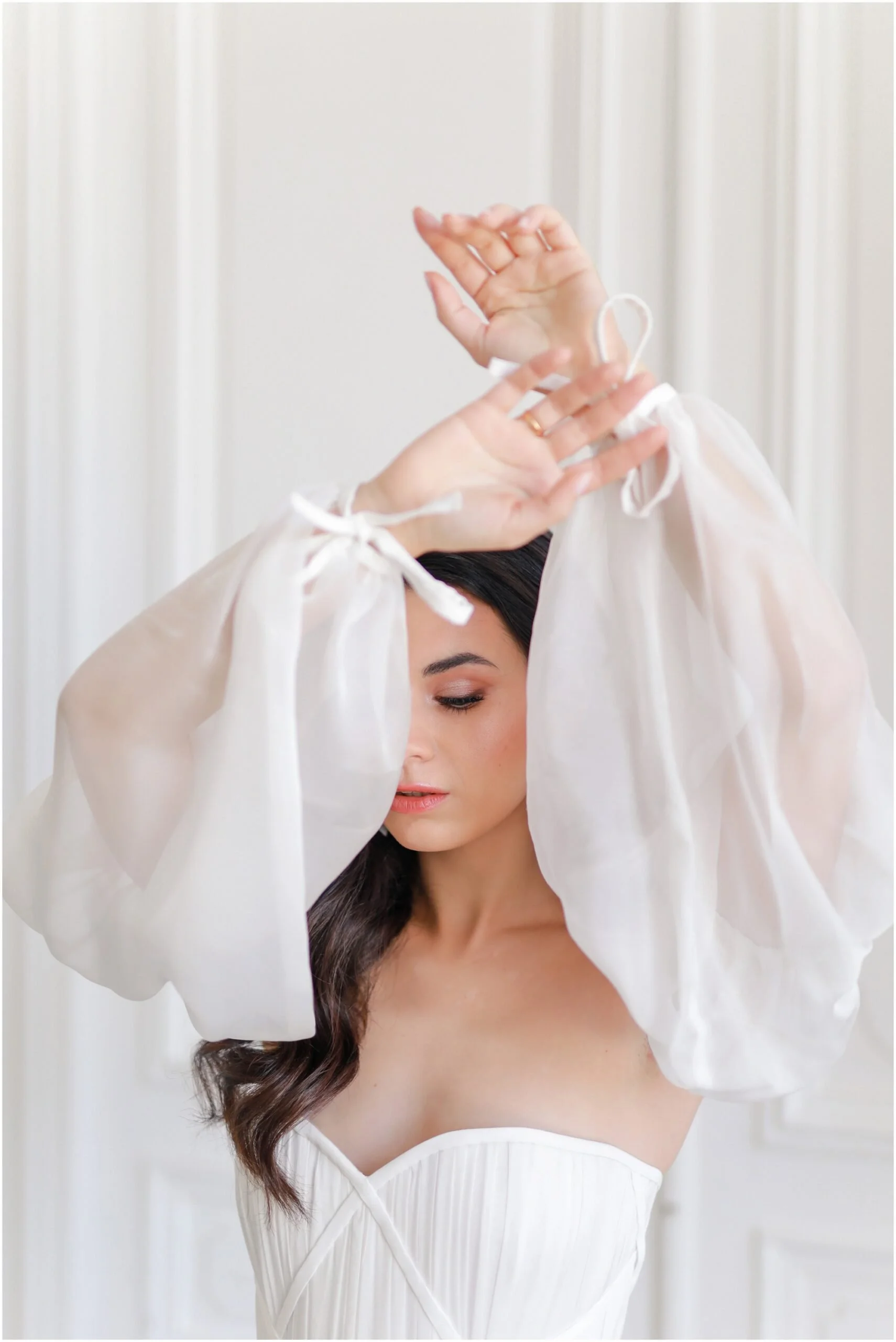 Une femme portant une robe blanche, dont les cheveux longs et bouclés tombent d'un côté, se tient devant un rideau blanc. Elle ajuste ses bras levés, portant des bracelets fins, dans un environnement lumineux et épuré.