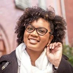 Marie-Michelle P. souriante portant des lunettes, avec des cheveux bouclés, portant une écharpe blanche et un manteau noir, dehors.