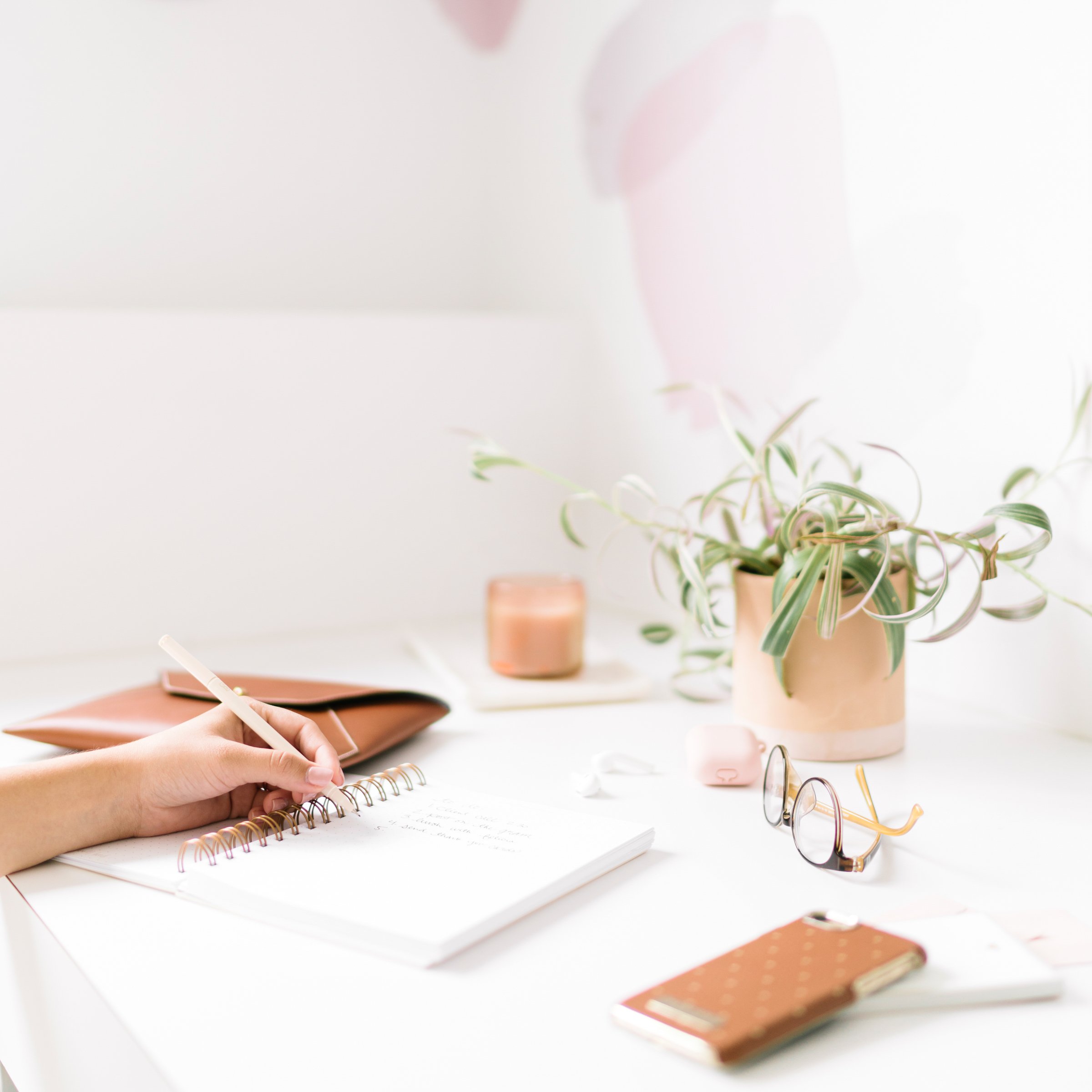 Une personne écrit dans un carnet blanc avec un stylo, sur une table blanche. À côté, il y a une paire de lunettes, un portefeuille gris et une montre. En arrière-plan, il y a plusieurs pots avec des plantes, notamment une grande avec des feuilles striées vertes et blanches. La scène est lumineuse et minimaliste.