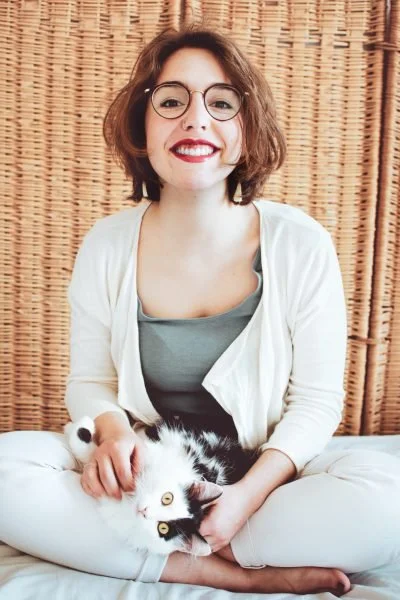 Marie B. assise en tailleur, tenant un chat noir et blanc, devant un fond en bois tressé.