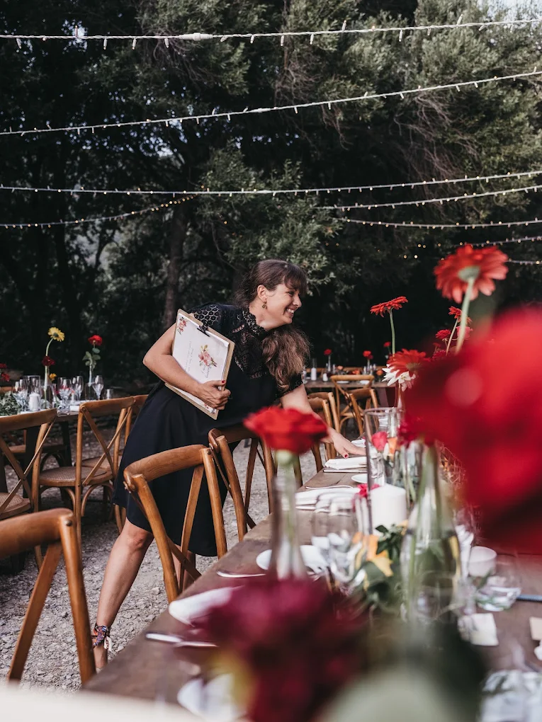 Une femme souriante, portant une robe noire, tenant un carnet ou un menu, se trouve près d'une longue table décorée de fleurs rouges et blanches, dans un cadre extérieur avec des arbres et des guirlandes lumineuses.