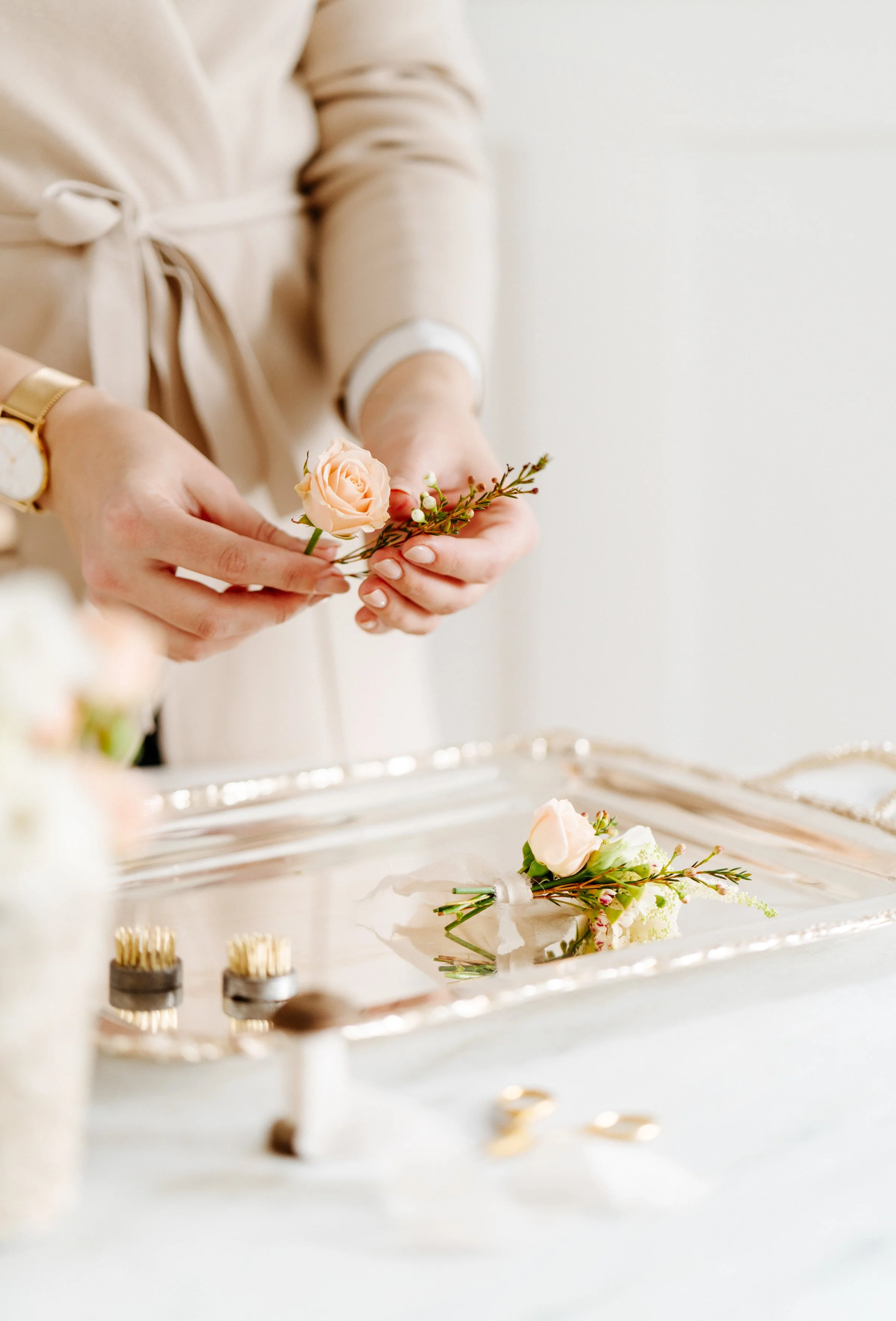 Une personne en robe beige prépare une boutonnière avec une petite rose crème et des feuillages, sur un plateau argenté avec d'autres boutonnières.