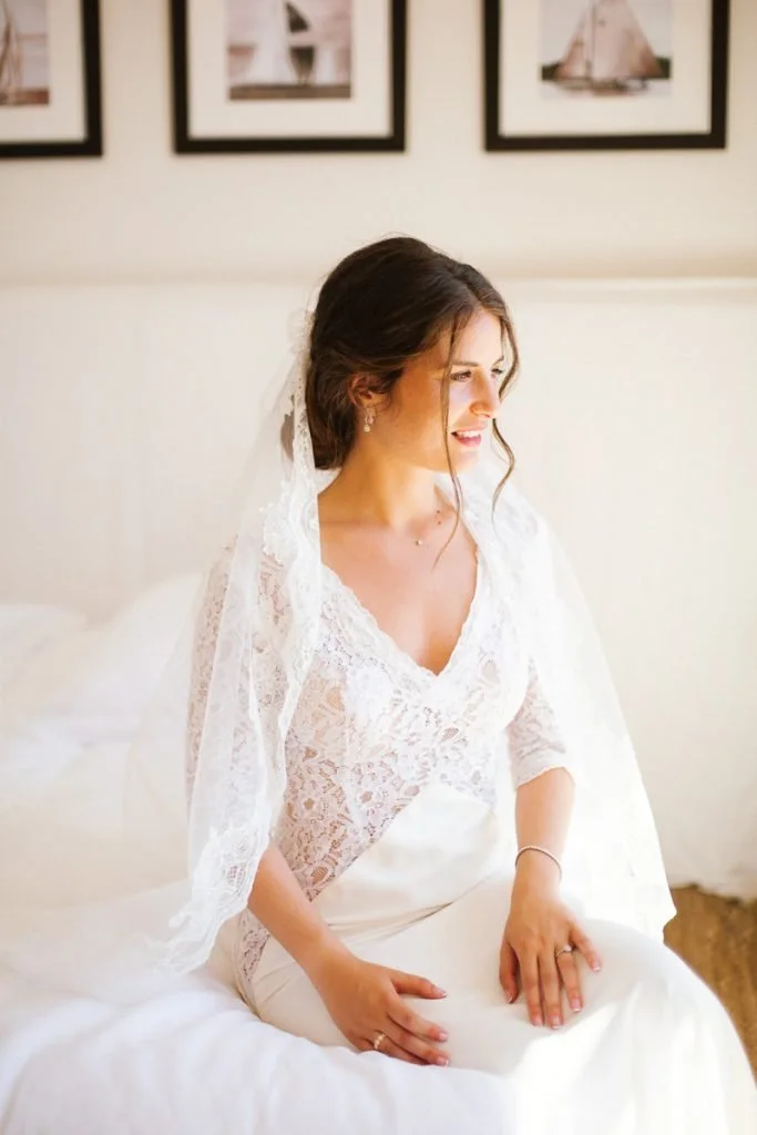 Une femme en robe de mariée en train de s'asseoir sur un lit, souriant en regardant vers la droite, dans une pièce décorée avec des photos encadrées sur le mur.