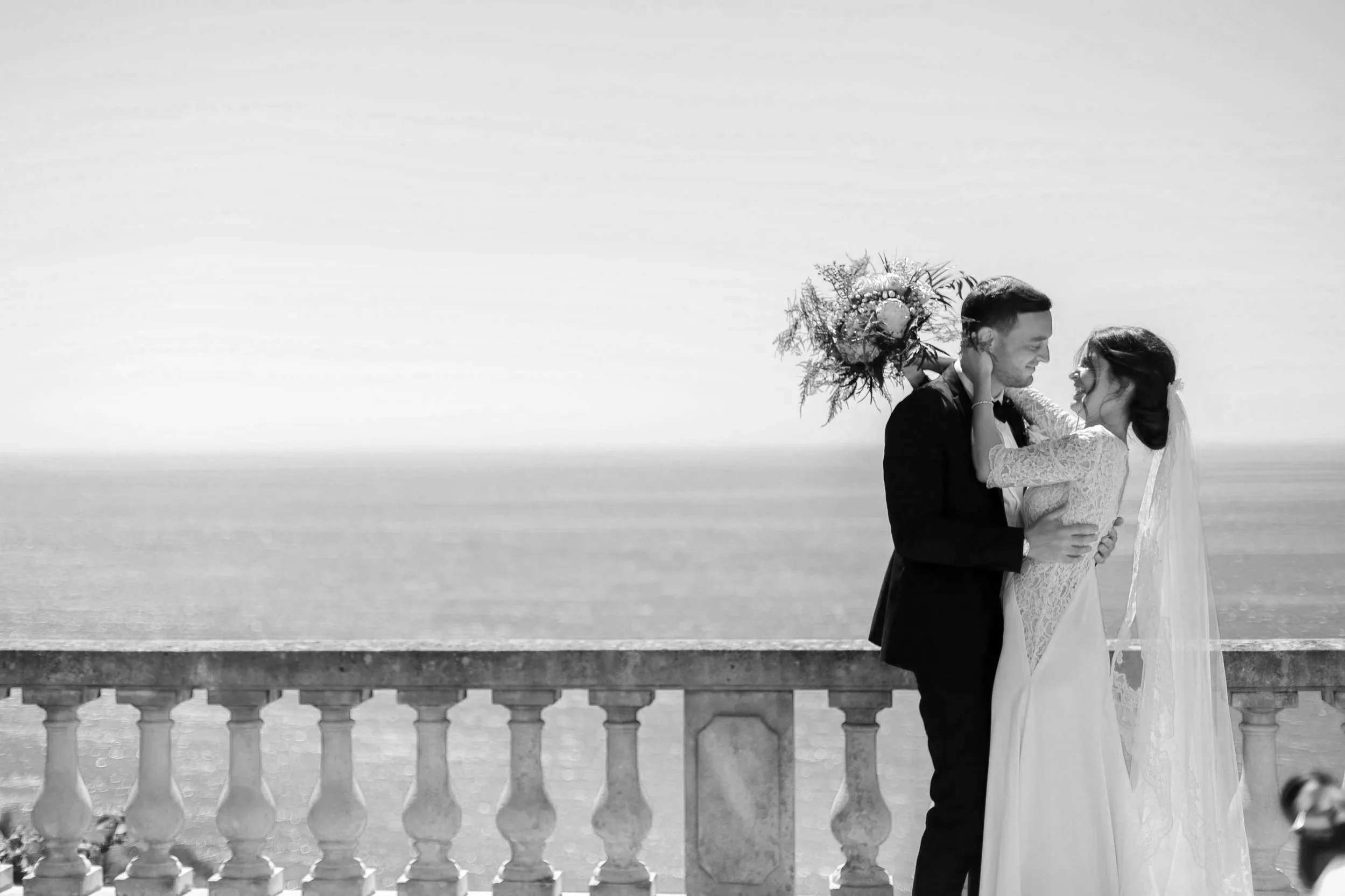Un couple en costume de mariage, souriant et se regardant, dans un décor en bord de mer, avec une balustrade en pierre.