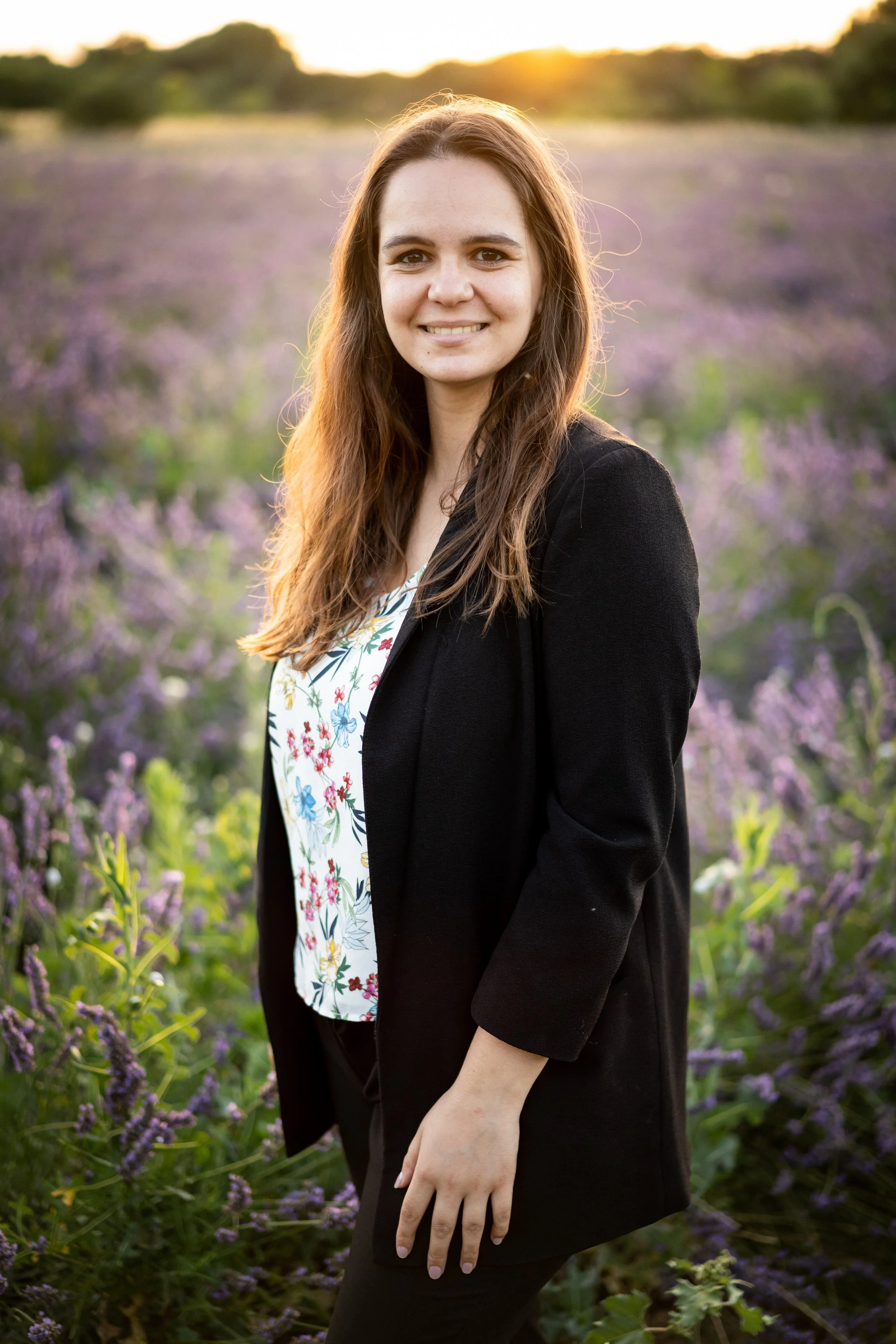 Chloé B. souriante avec des cheveux longs marron, portant un blazer noir et une robe à motifs floraux, debout dans un champ de lavande au coucher du soleil.