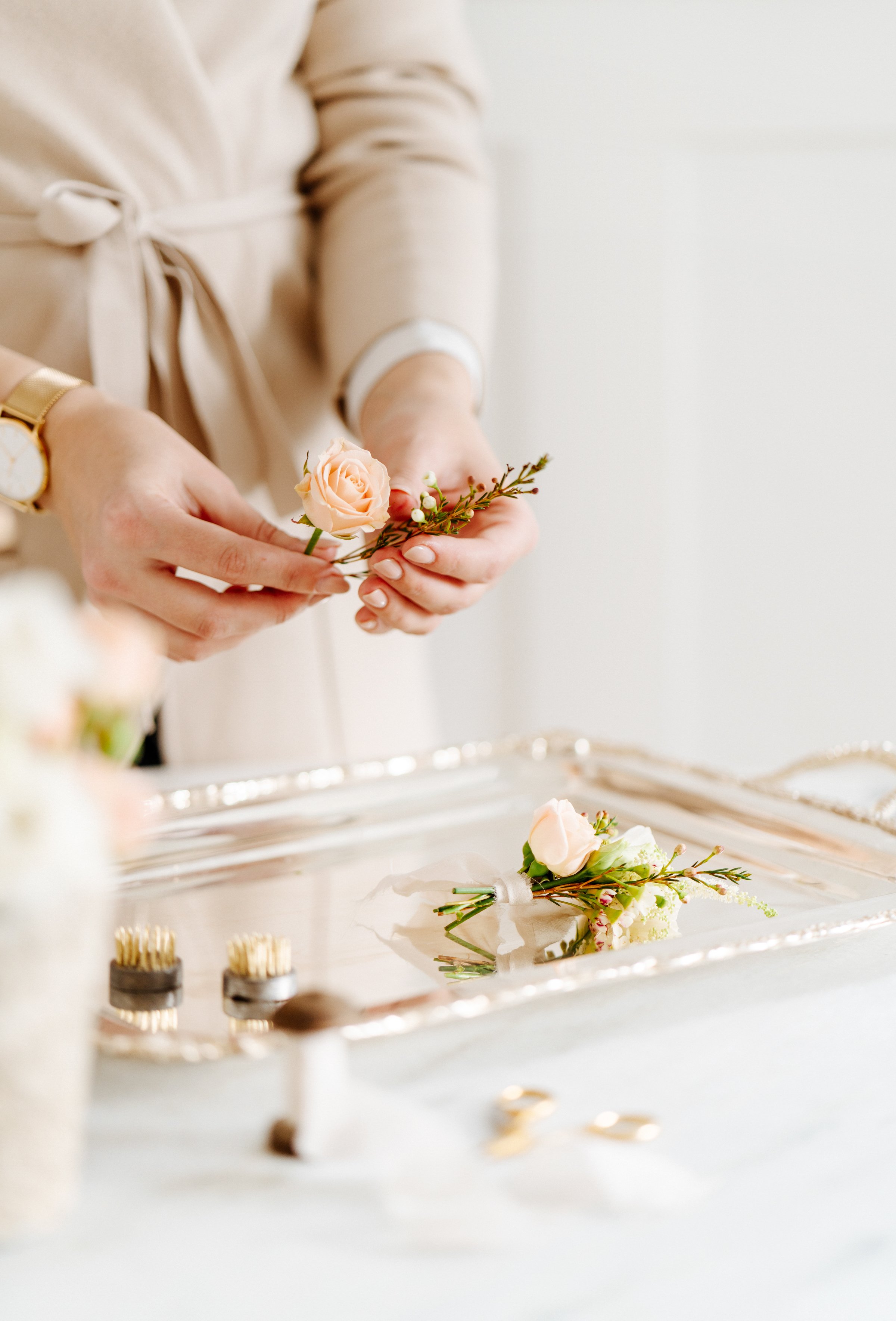Une personne arrange une petite boutonnière avec une rose pâle et des brindilles, posée sur une surface de couleur claire. D'autres boutonnières en fleurs sont visibles sur une surface argentée.