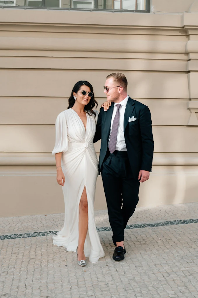 Un couple habillé élégamment, la femme en robe blanche et le homme en costume noir, marchant côte à côte devant un mur beige.