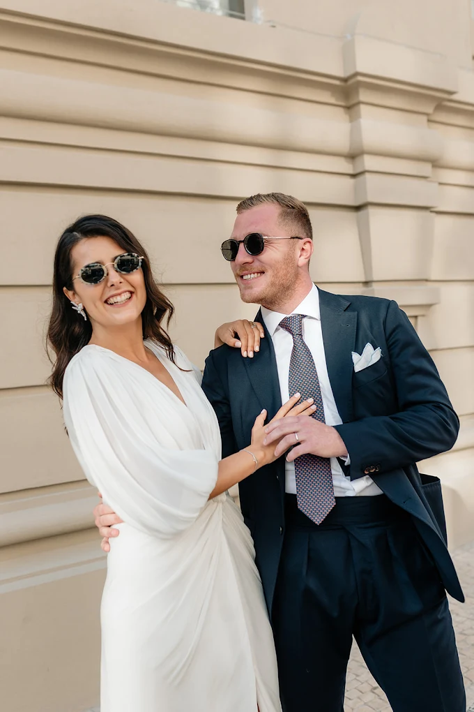 Un couple habillé en vêtements élégants, la femme en robe blanche et l'homme en costume sombre, souriants et portant des lunettes de soleil, se tenant devant un bâtiment en pierre.