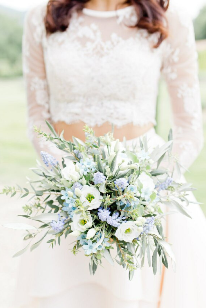 Femme portant une robe de mariage en dentelle blanche, tenant un bouquet de fleurs blanches, bleues et vertes, dans un cadre extérieur verdoyant.