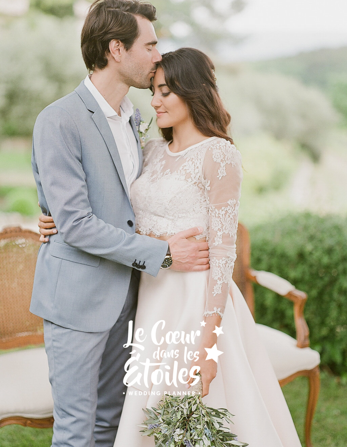 Un couple lors d'une cérémonie de mariage. Le marié, en costume gris clair, embrasse tendrement la mariée qui porte une robe blanche en dentelle. La mariée tient un bouquet de fleurs vertes et violettes. L'arrière-plan est un jardin avec des chaises et des plantes. Le texte sur l'image dit "Le Cœur dans les Étoiles, Wedding Planner".