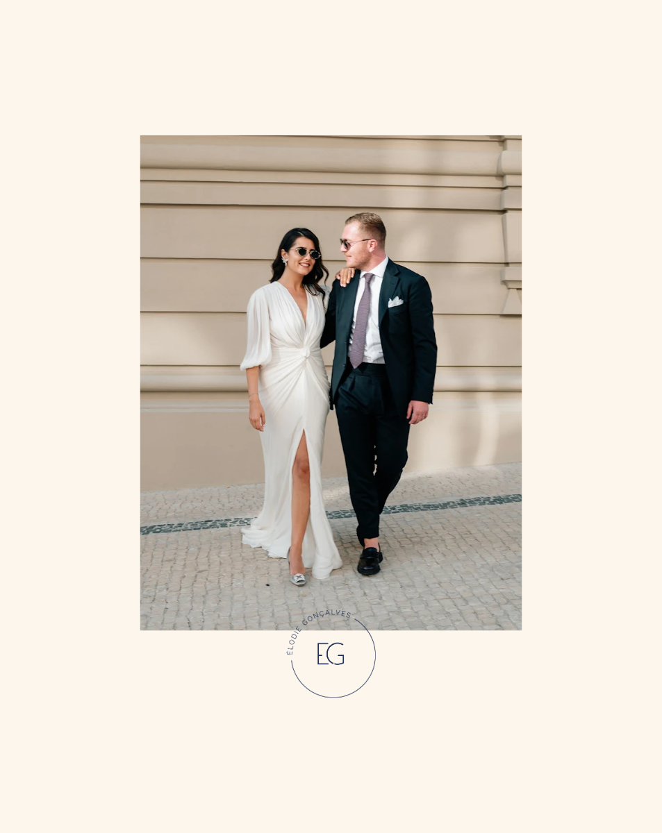 Un couple en tenue élégante marchant côte à côte devant un mur beige, la femme porte une robe blanche longue avec une fente et des lunettes de soleil, l'homme porte un costume sombre avec une cravate, ils sourient et semblent liés par une relation amoureuse ou amicale.