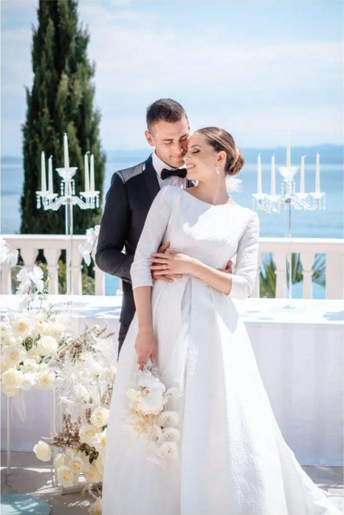Un couple marié posant lors d'un mariage en extérieur avec vue sur l'eau, décor avec chandeliers et fleurs blanches.