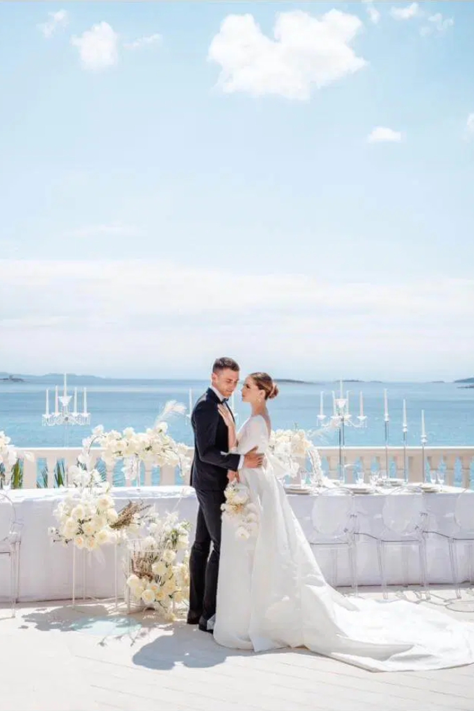 Un couple de mariés se tenant par la taille lors de leur mariage sur une terrasse près de la mer, décorée de fleurs blanches et de chandeliers, avec un ciel bleu en arrière-plan.