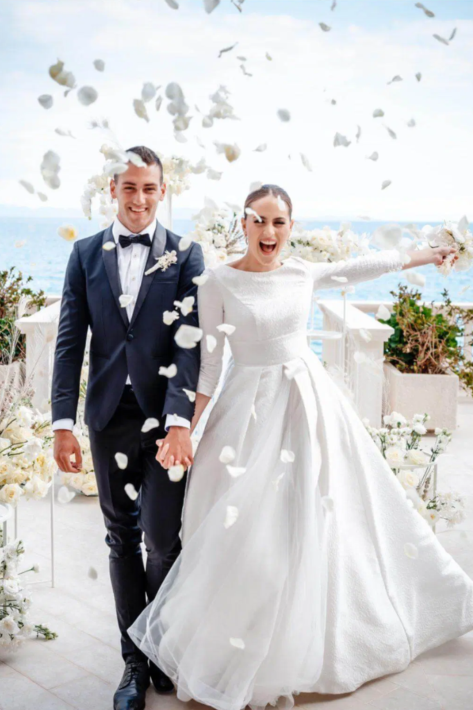 un couple de mariés souriants, mains jointes, entourés de fleurs blanches, lors de leur mariage en plein air avec vue sur la mer, lors d'une cérémonie joyeuse