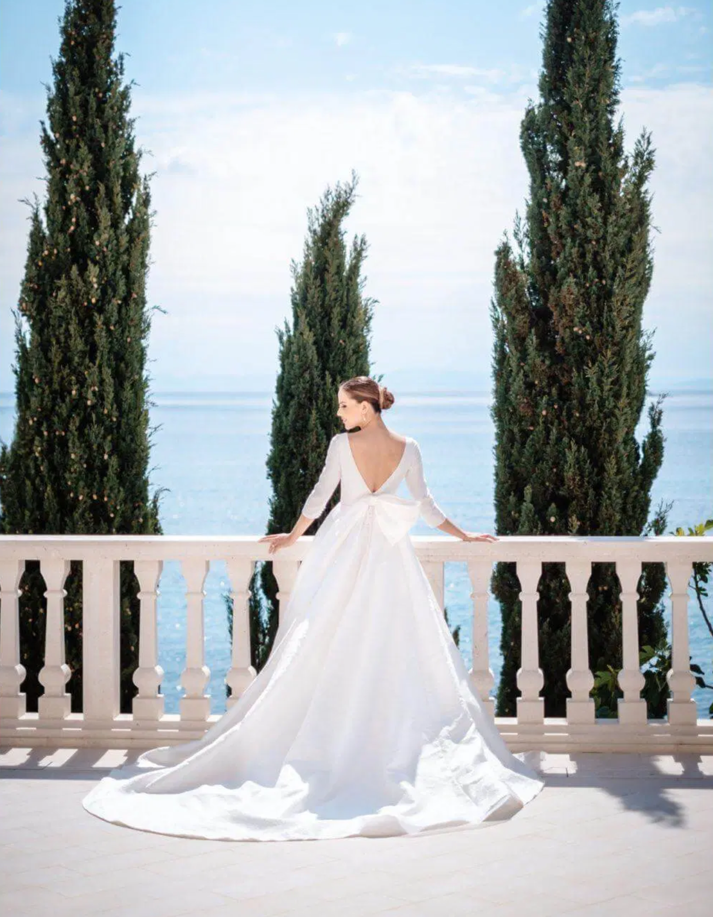 Une femme en robe de mariage blanche sur une terrasse avec vue sur la mer, entourée de trois grands arbres vert foncé.