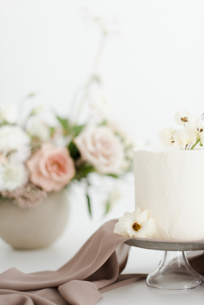 Gâteau blanc décoré de fleurs blanches et crème, avec un arrangement floral de roses et autres fleurs en arrière-plan.