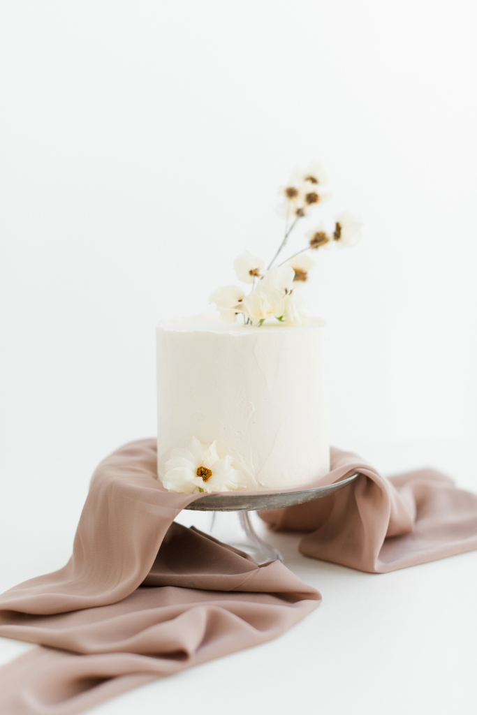 Gâteau blanc minimaliste décoré de fleurs blanches, posé sur une draperie en tissu beige