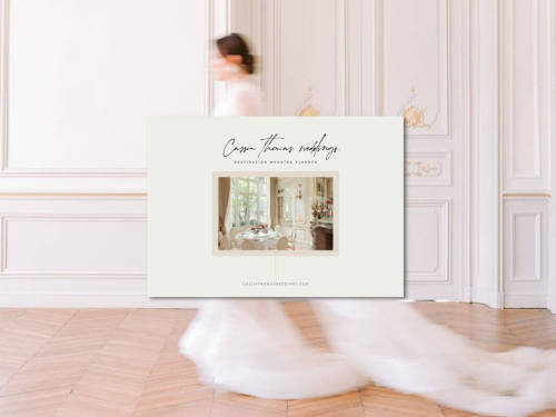 Une femme en robe de mariée blanc marche dans une pièce élégante avec des murs ornés et un parquet en bois, passant devant une grande affiche promotionnelle pour un wedding planner.