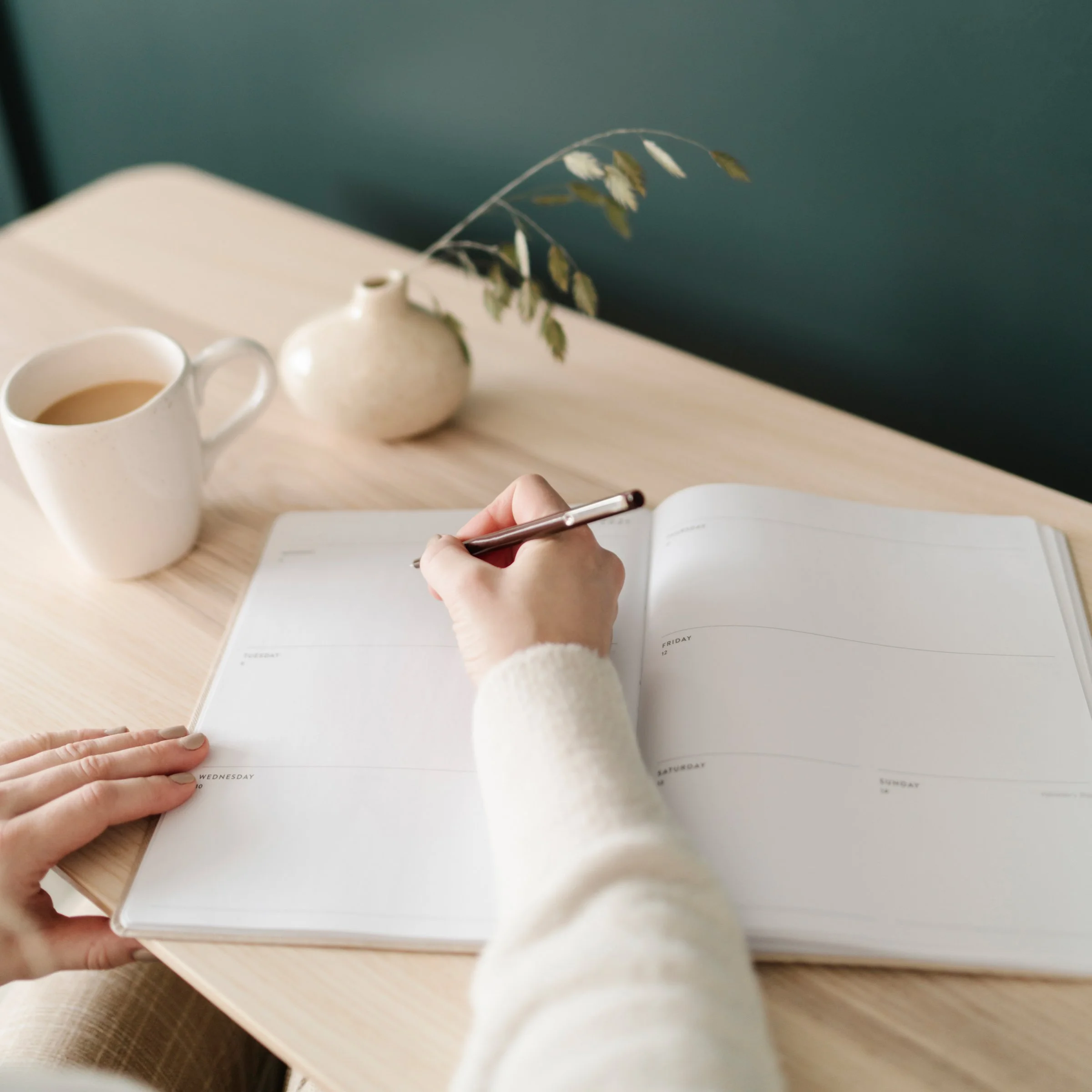Une main écrivant dans un diary ouvert posé sur une table en bois, avec une tasse de café et une petite cruche blanche à côté, en fond un mur vert. L'image est floue.