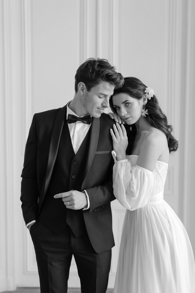Un homme en costume noir avec un nœud papillon et une femme en robe blanche formelle posent ensemble, le fond d'une pièce élégante.