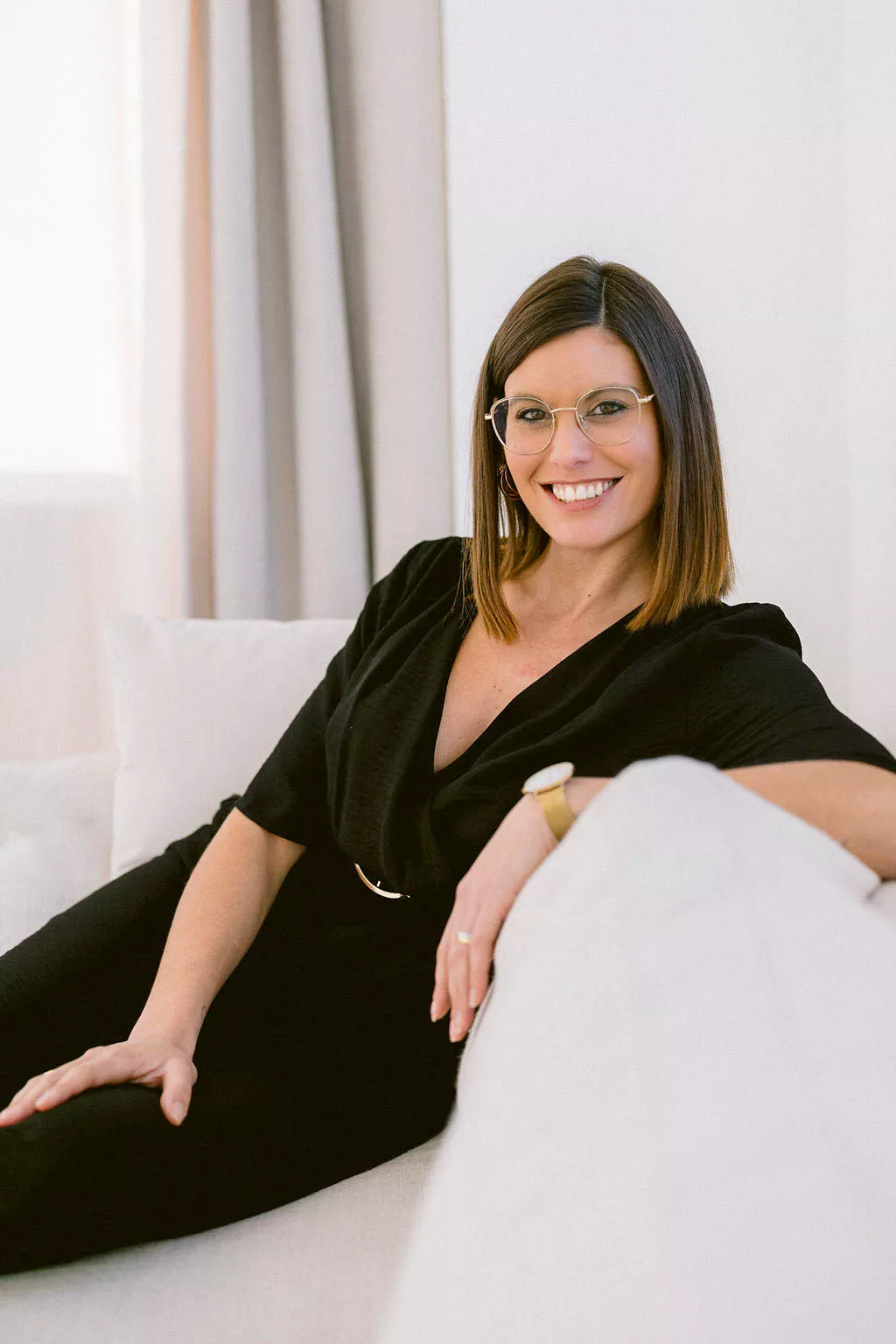 Justine Huette souriante avec des lunettes, assise sur un canapé blanc, portant une robe noire et un bracelet doré.