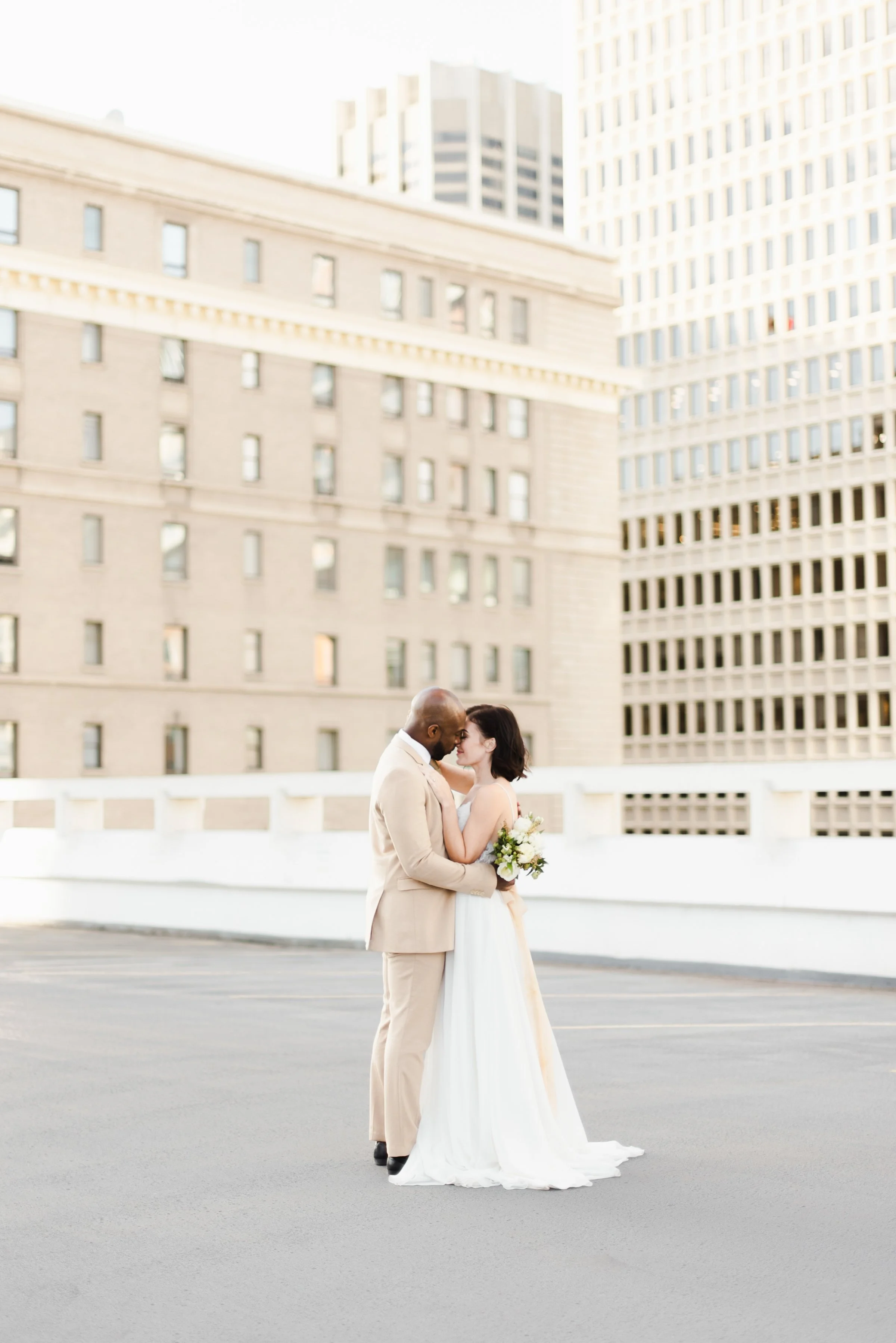 Un couple en mariage se tient dans une ville moderne, le homme habillé en costume beige et la femme en robe de mariée blanche, s'enlaçant tendrement. La femme tient un bouquet de fleurs. En arrière-plan, il y a des bâtiments de bureaux en pierre claire et en verre, avec un ciel ensoleillé.