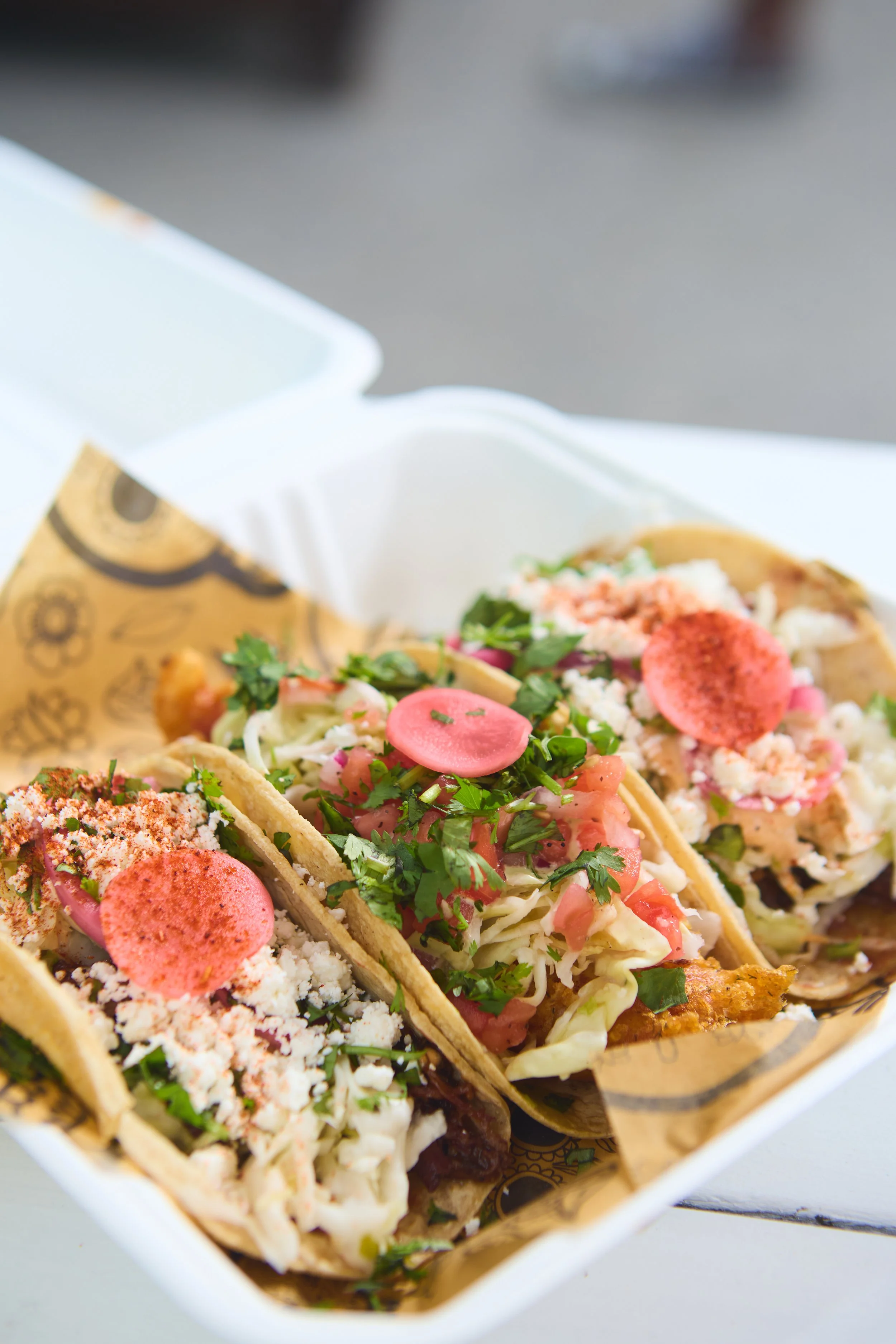 Three tacos with various toppings including cilantro, chopped onions, diced tomatoes, and pink radishes, served in a paper tray.