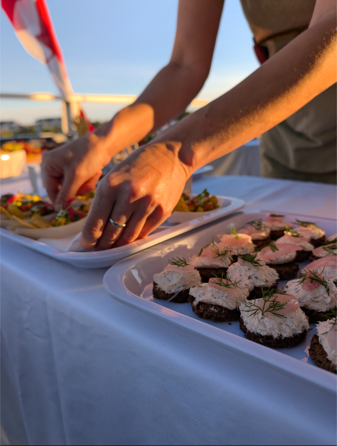 Person arranging small appetizers on a white tablecloth at an outdoor event during sunset, with a selection of food on plates.