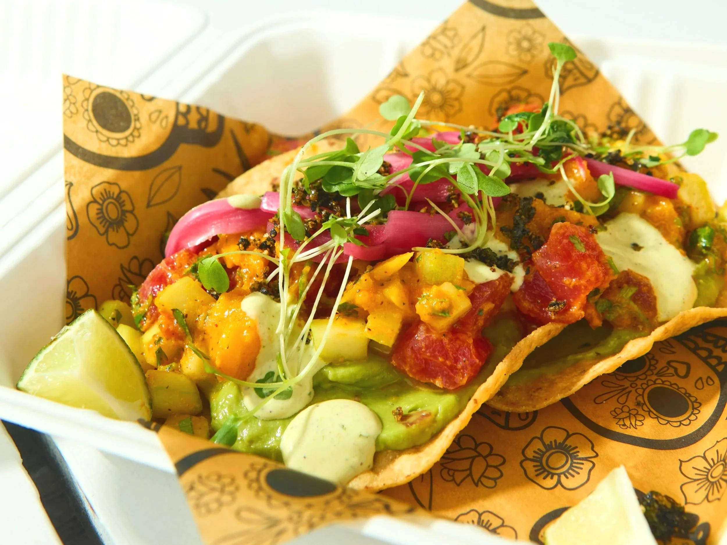 Close-up of a taco with various colorful toppings including microgreens, pickled onions, and diced vegetables, served on a paper-lined tray with lime wedges.
