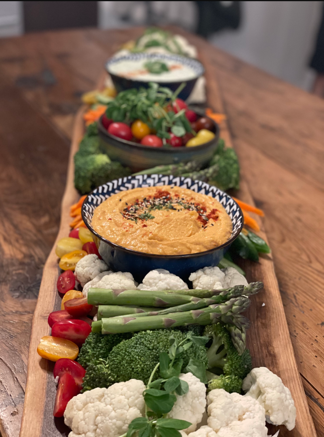 A long wooden serving board with bowls of dip and an assortment of fresh vegetables including cherry tomatoes, cauliflower, broccoli, asparagus, carrots, and mixed greens.