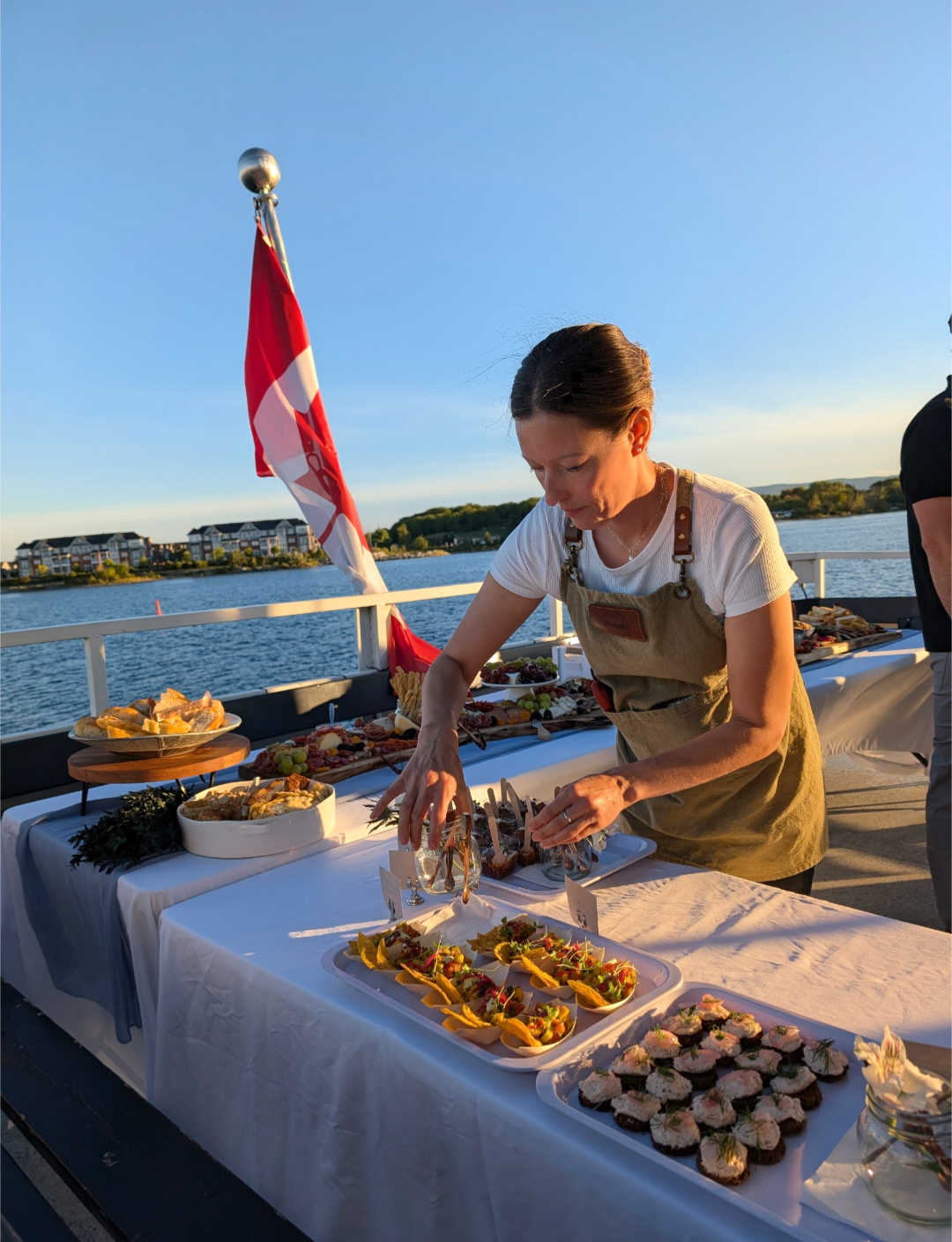 A woman in a white shirt and beige apron arranging food on a table set on a boat, with water and buildings in the background during sunset.