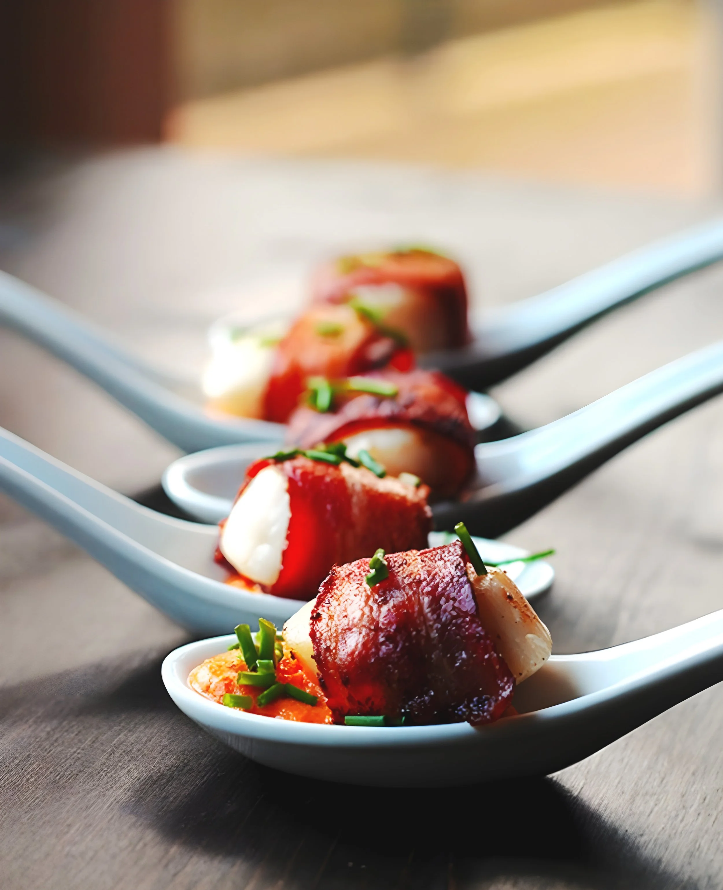 Close-up of appetizers with bacon and cheese garnished with chopped green herbs on white ceramic spoons arranged on a dark wooden surface.