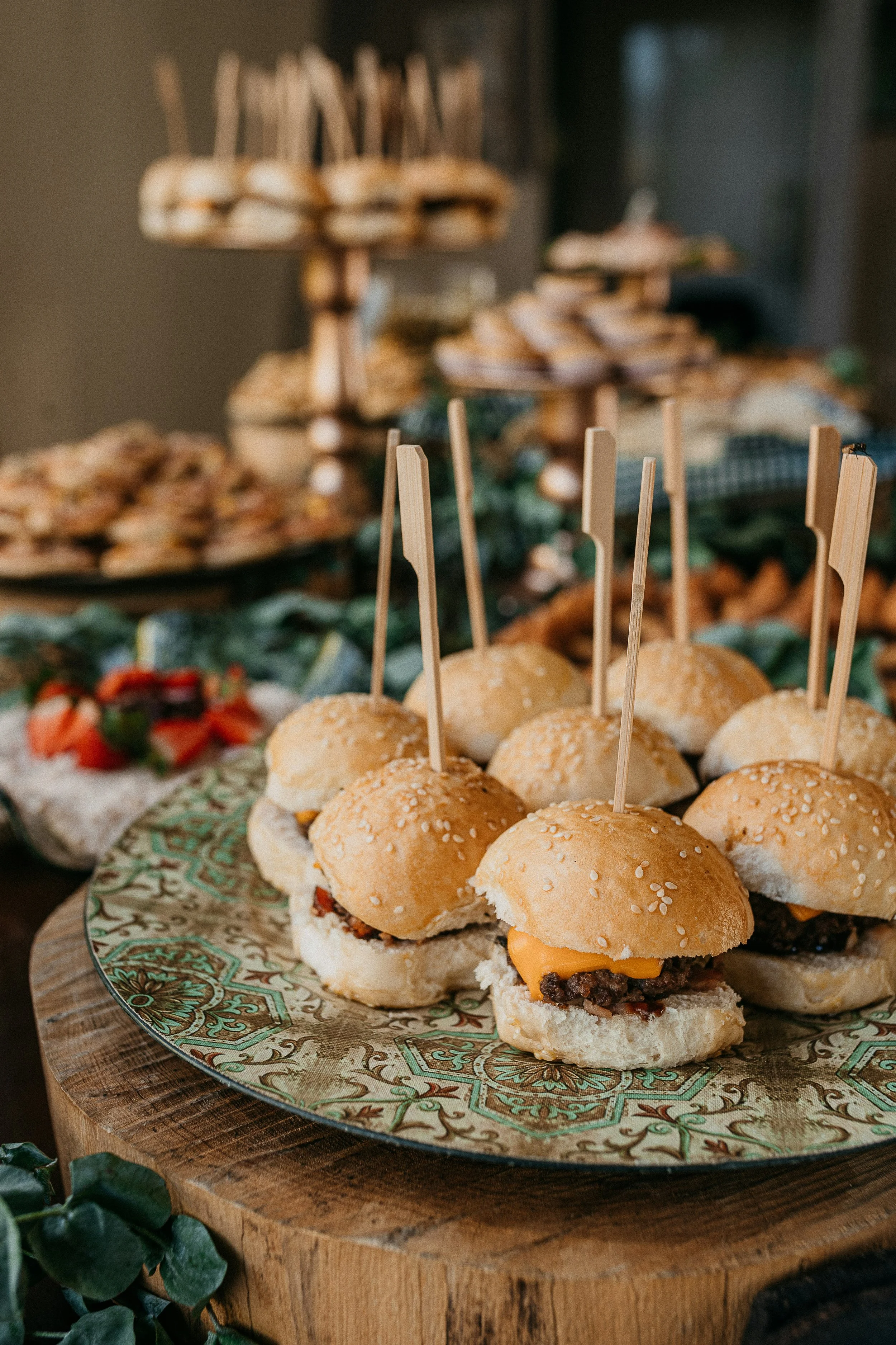 Close-up of mini cheeseburgers with sesame seed buns and small wooden skewers, arranged on a decorative plate on a wooden serving board, with a platter of sliders in the background.