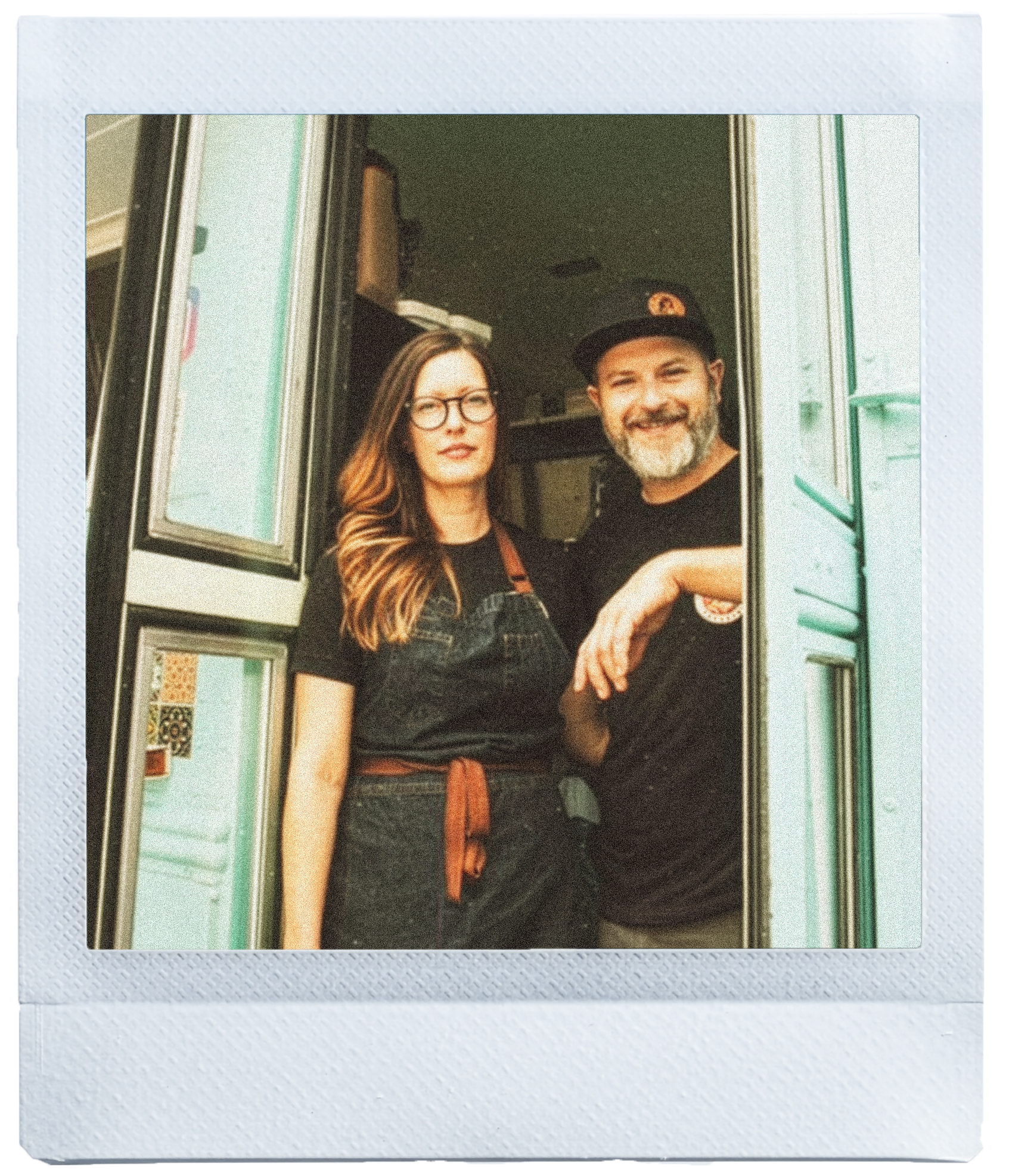 Two people, a woman with glasses and a man with a beard wearing a baseball cap, standing in a window while taking a selfie.