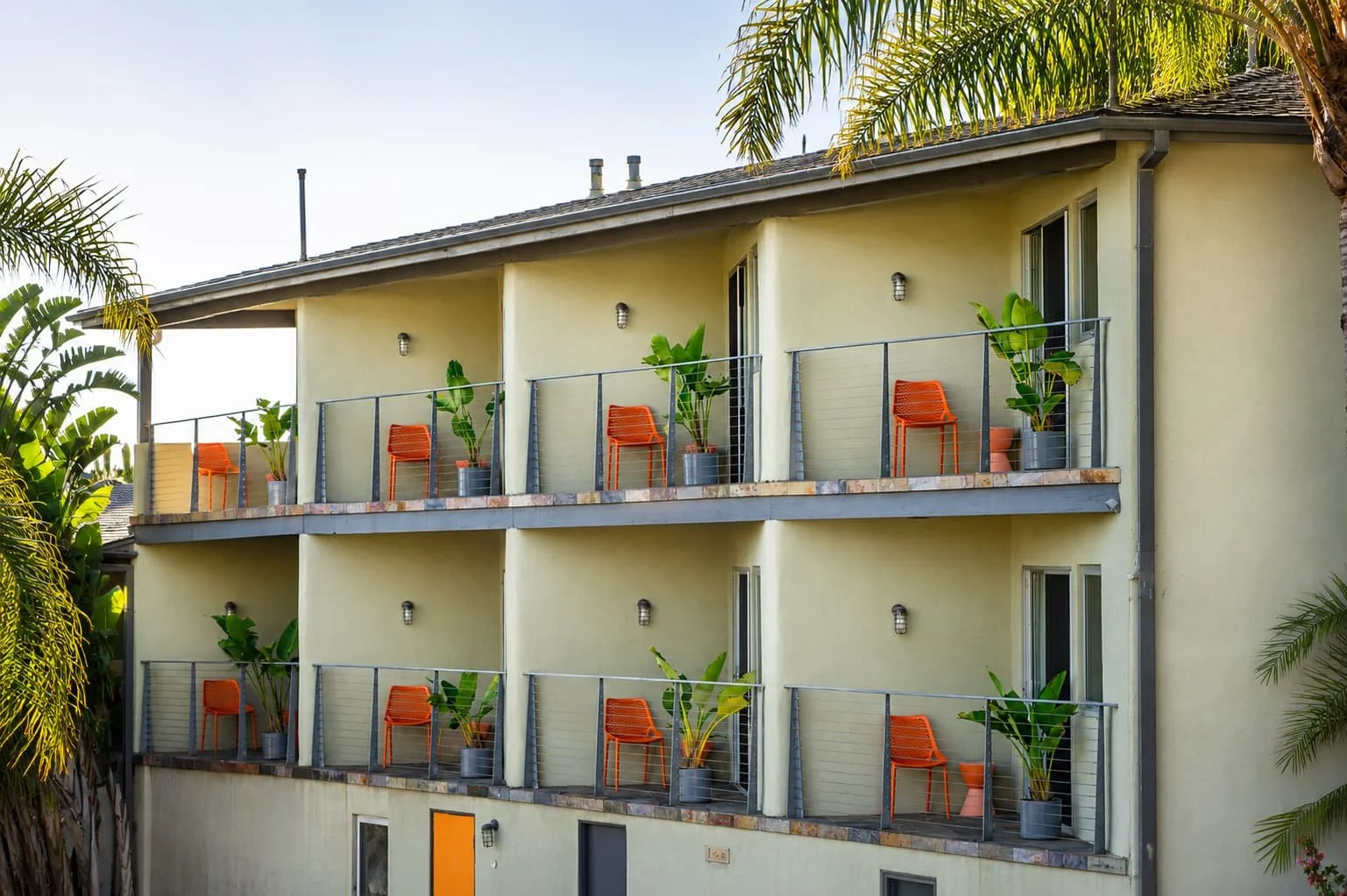 Three-story Malibu Beach hotel with balconies, orange chairs, potted plants, all surrounded by palm trees.