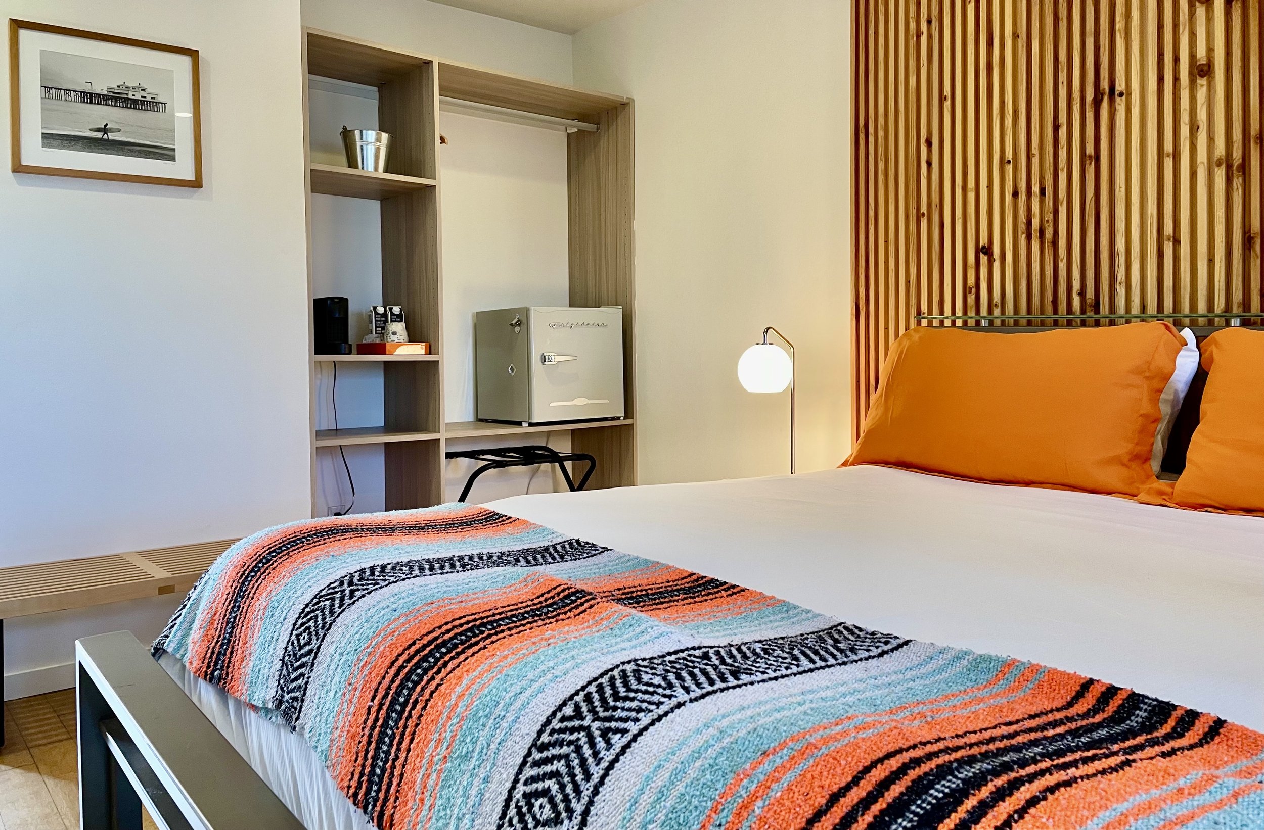 The Surfers Spot room featuring a slatted bed frame, a mexican blanket on the top of the bedding, an art piece by a local artist, and the closet which has the amenities like the small fridge and Nespresso machine.