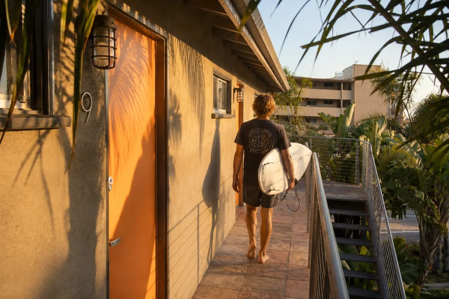 Barefoot person with a surfboard walks an outdoor corridor toward stairs, near an orange door marked 9 at a surfers hotel.
