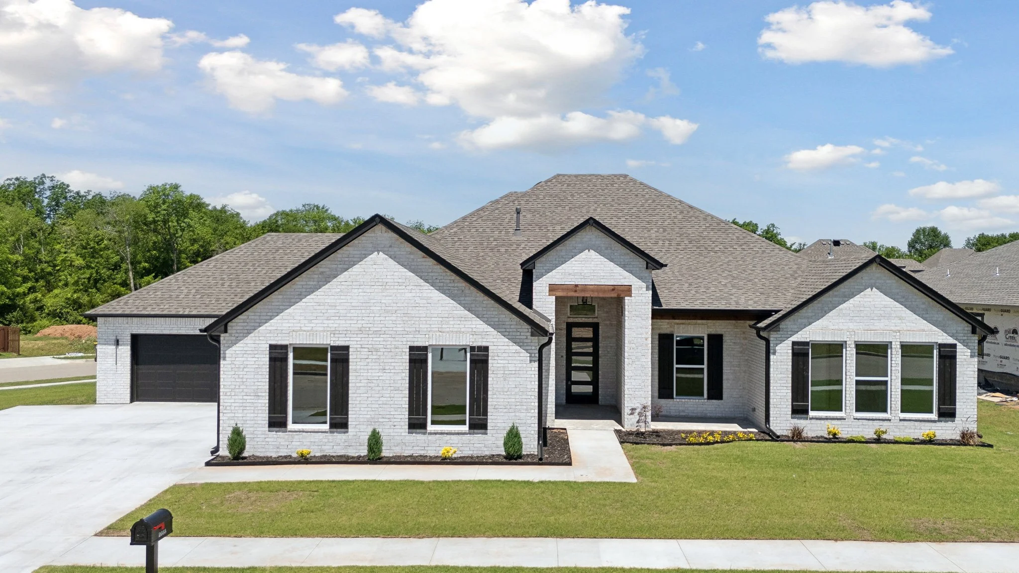 Newly built white brick house with black trim and a gray shingled roof, front yard with green grass and small bushes, concrete driveway and mailbox, under a partly cloudy sky.
