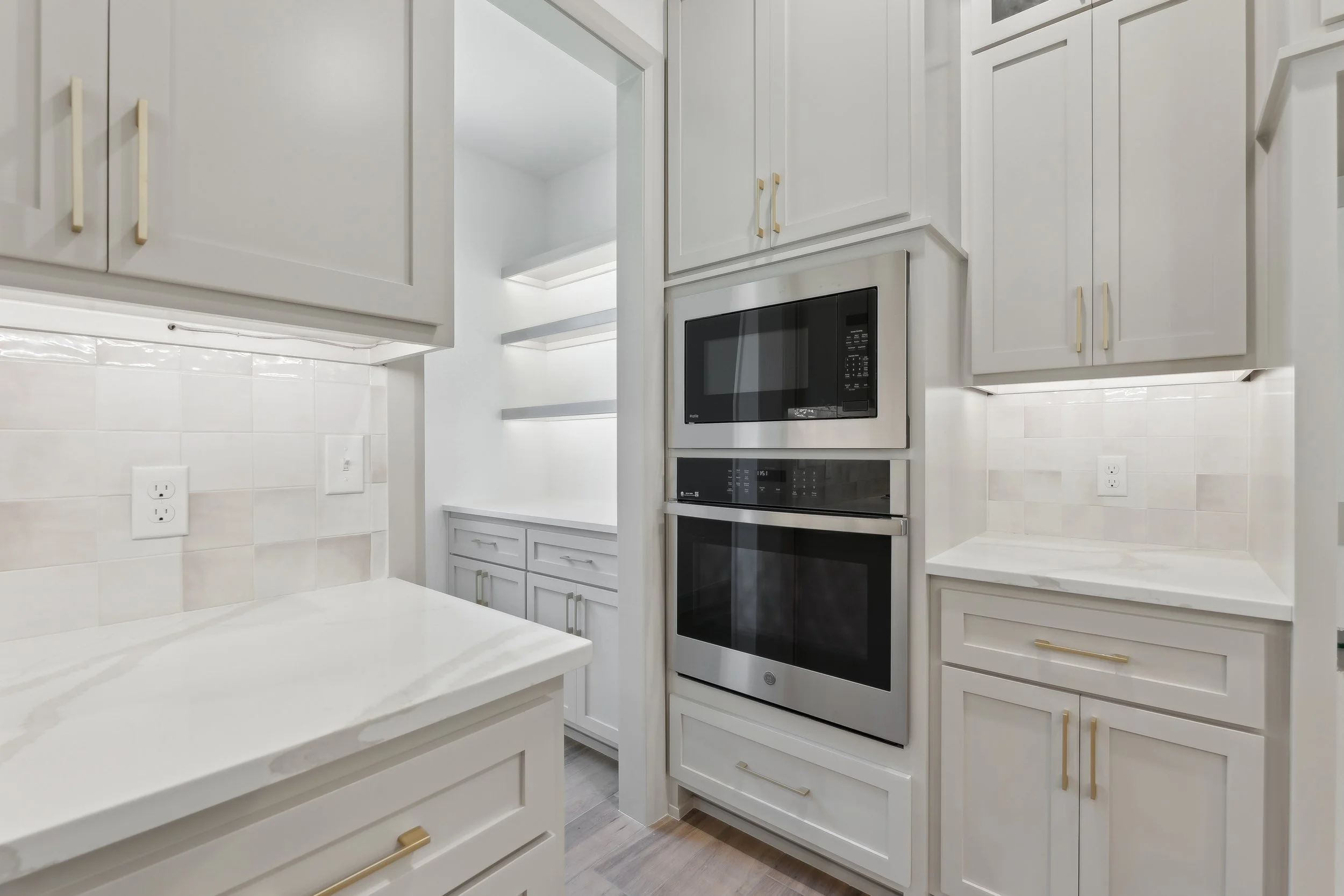 A modern white kitchen with built-in microwave and oven, white cabinets with gold handles, white countertops, and a gray wooden floor.