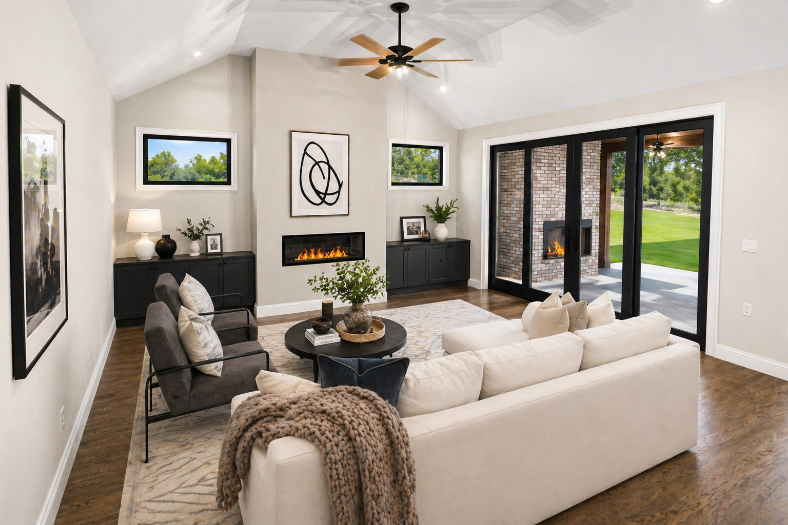 A modern living room with a beige sectional sofa, a black round coffee table, and two black armchairs. There are two black cabinets with decor and a fireplace on a white wall. Large sliding glass doors lead to an outdoor patio with a brick fireplace and green backyard.