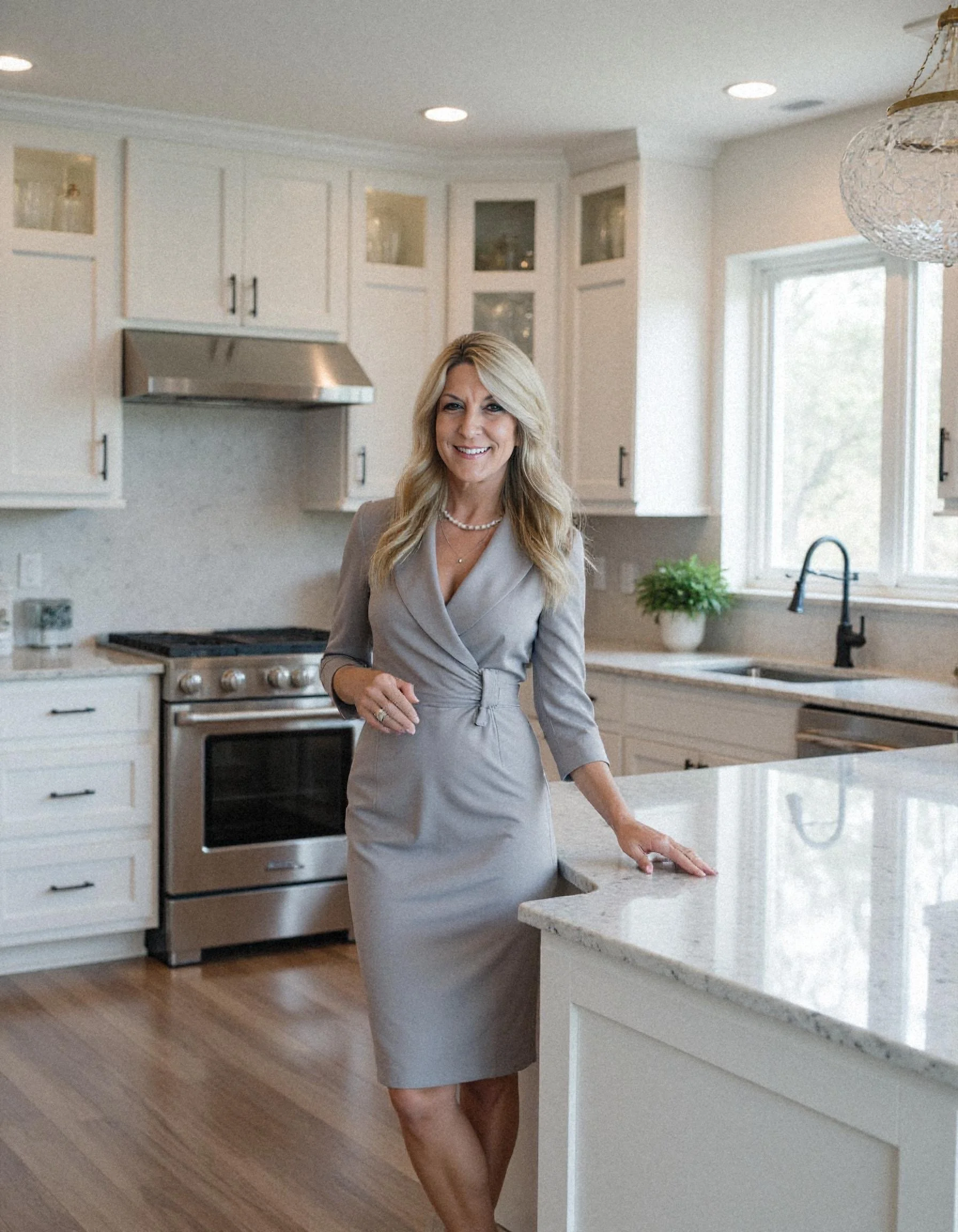 A woman in a gray dress standing in a modern, white kitchen with natural light coming in through a window, smiling at the camera.