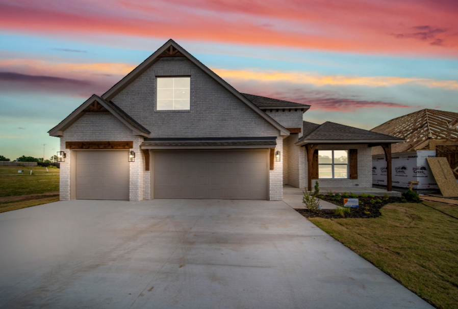 Newly constructed two-story house with a three-car garage, brick exterior, and front porch under construction, with a colorful sunset sky in the background.