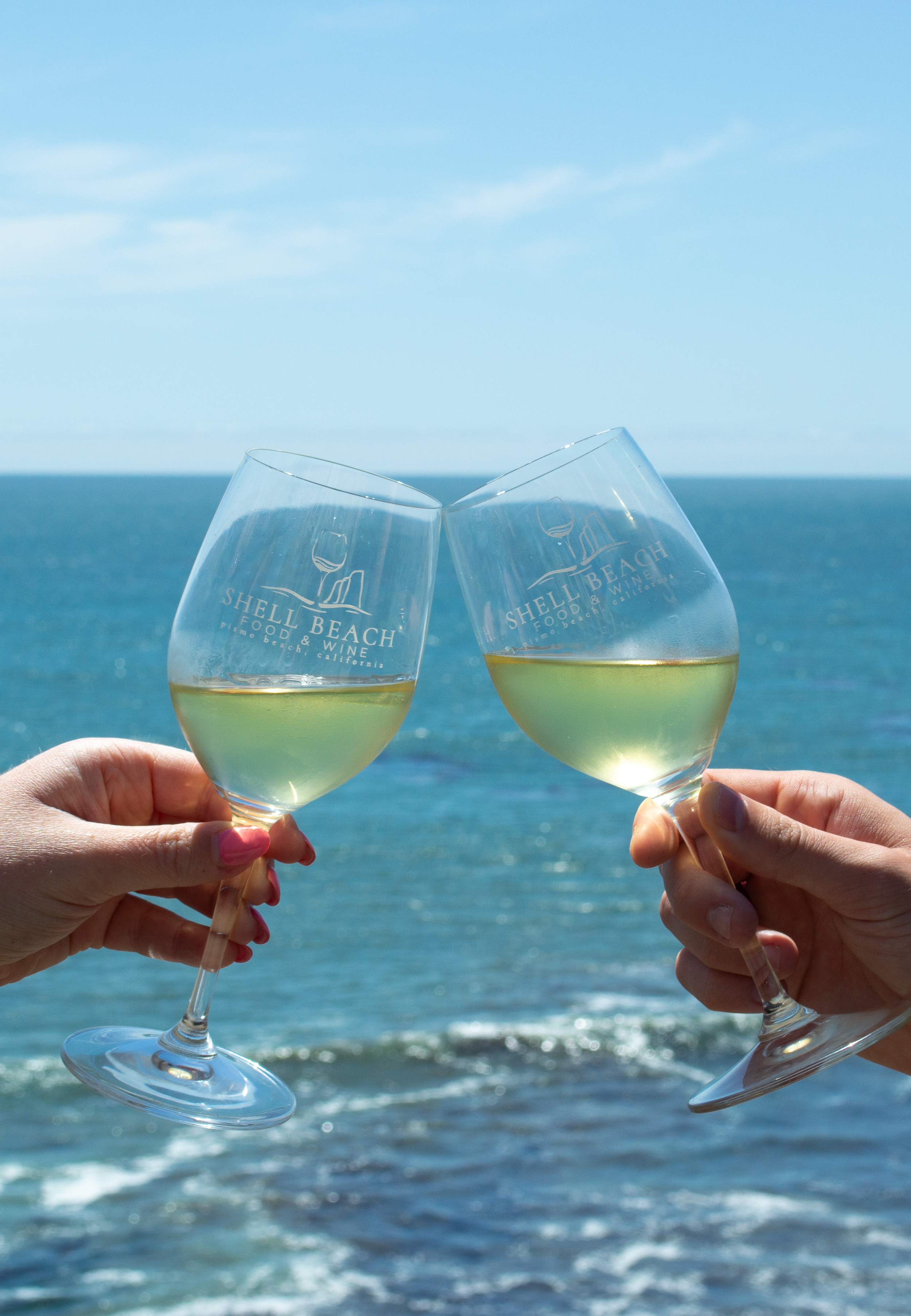 Two hands holding Shell Beach Food & Wine branded wine glasses clinking together against an ocean background with a blue sky at Dolphin Bay Resort & Spa in Pismo Beach, California. Celebrating central coast at this annual oceanfront tasting event.