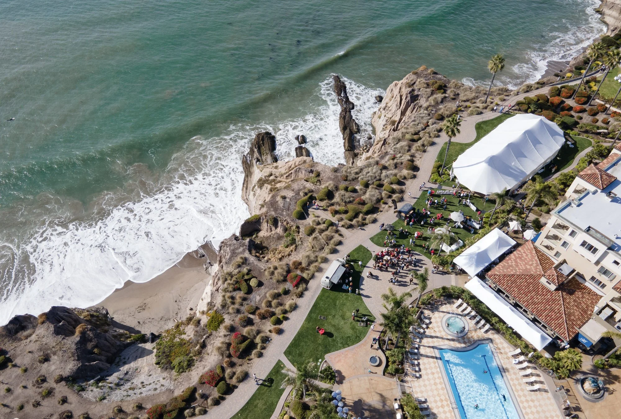 Aerial view of Shell Beach Food & Wine at Dolphin Bay Resort & Spa in Pismo Beach with a tented event space, a beach, and rocky cliffs with ocean waves, whiskey tastings, tequila tastings on the oceanfront lawn.