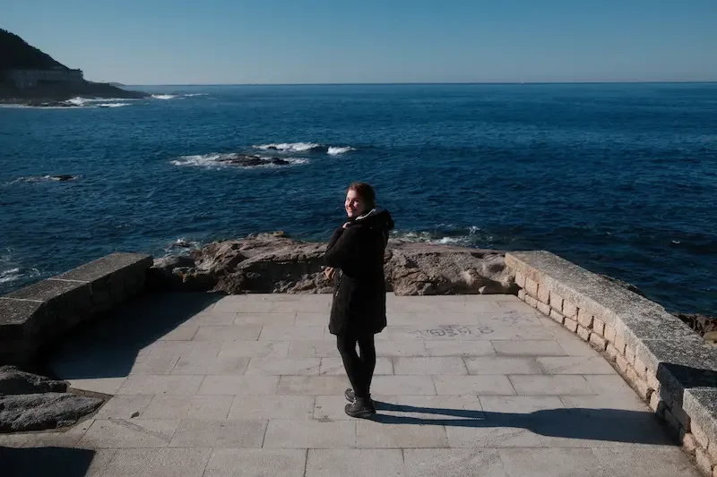 Teresa dos Reis standing on a paved viewing area by the ocean, with rocky shoreline in the background, on a sunny day.