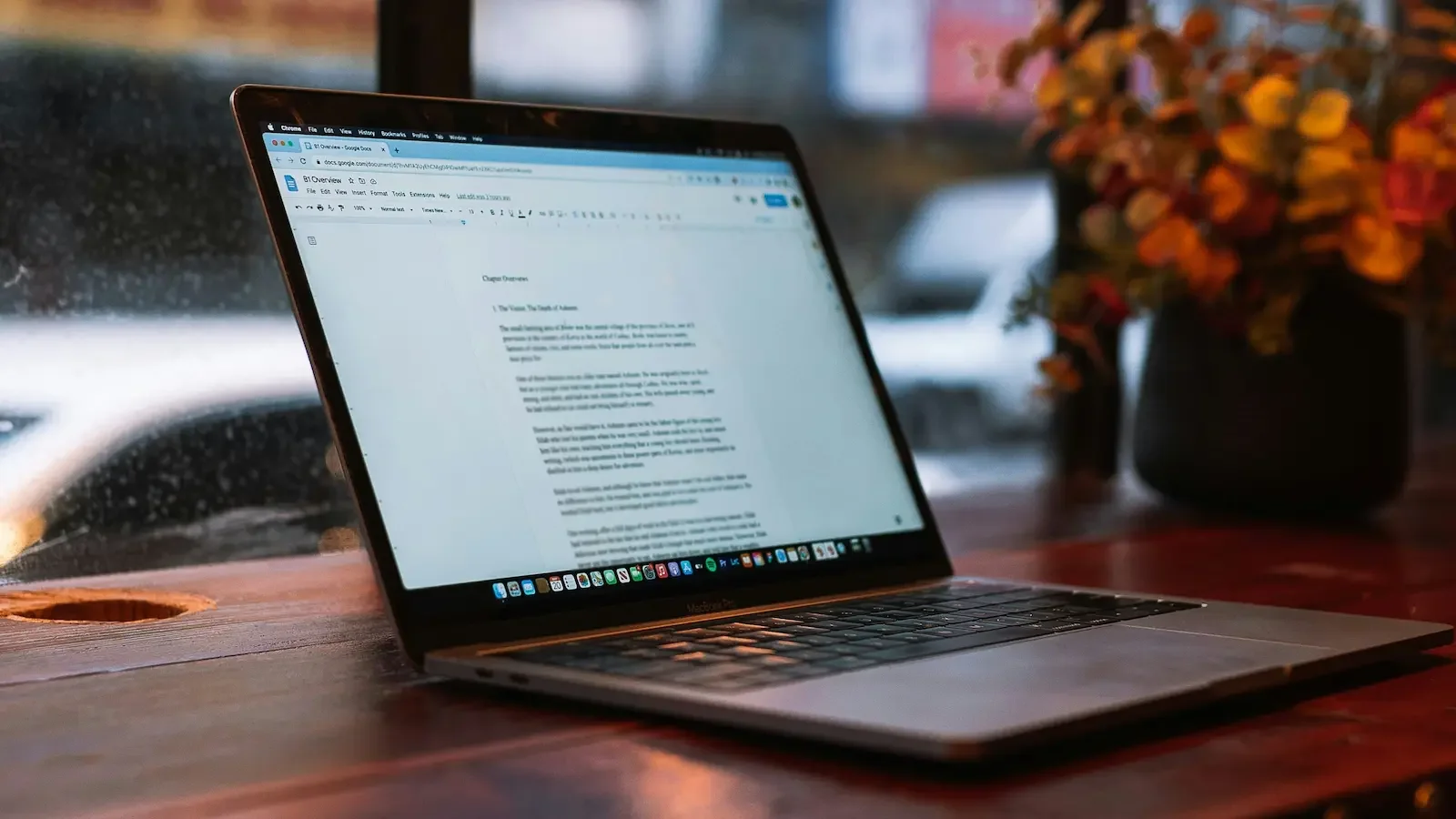 Open laptop on a wooden desk by a blurred rain-soaked window, with a potted plant reflecting golden light